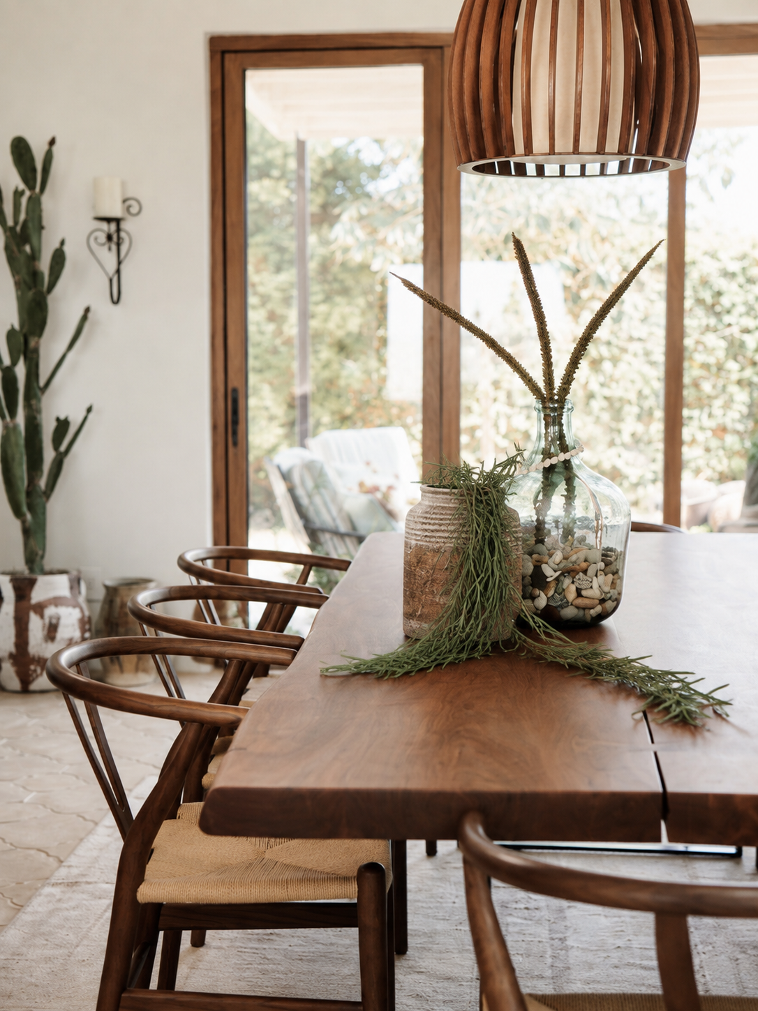 A rustic wooden dining table with matching chairs in a bright, modern dining room. A clear glass vase with dried grasses and greenery sits on the table, under a slatted wooden pendant light.