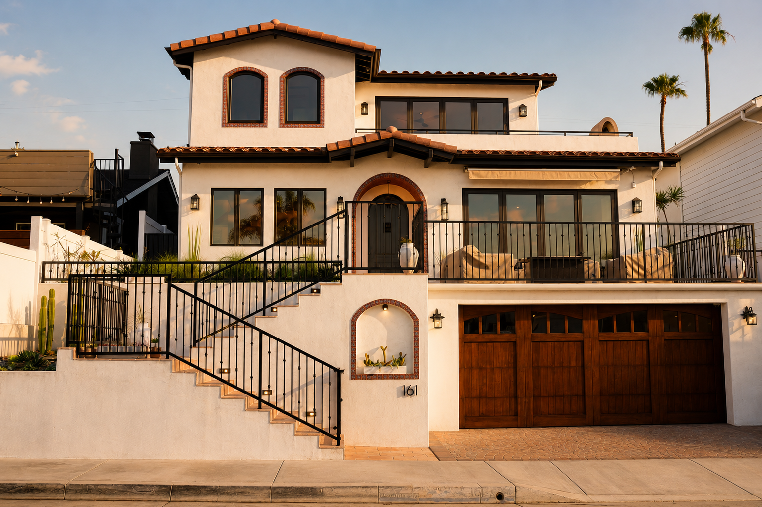 A two-story Hacienda-style house exterior with white stucco walls, terracotta roof tiles, black metal railings, a staircase, a garage door, and palm trees in the background.