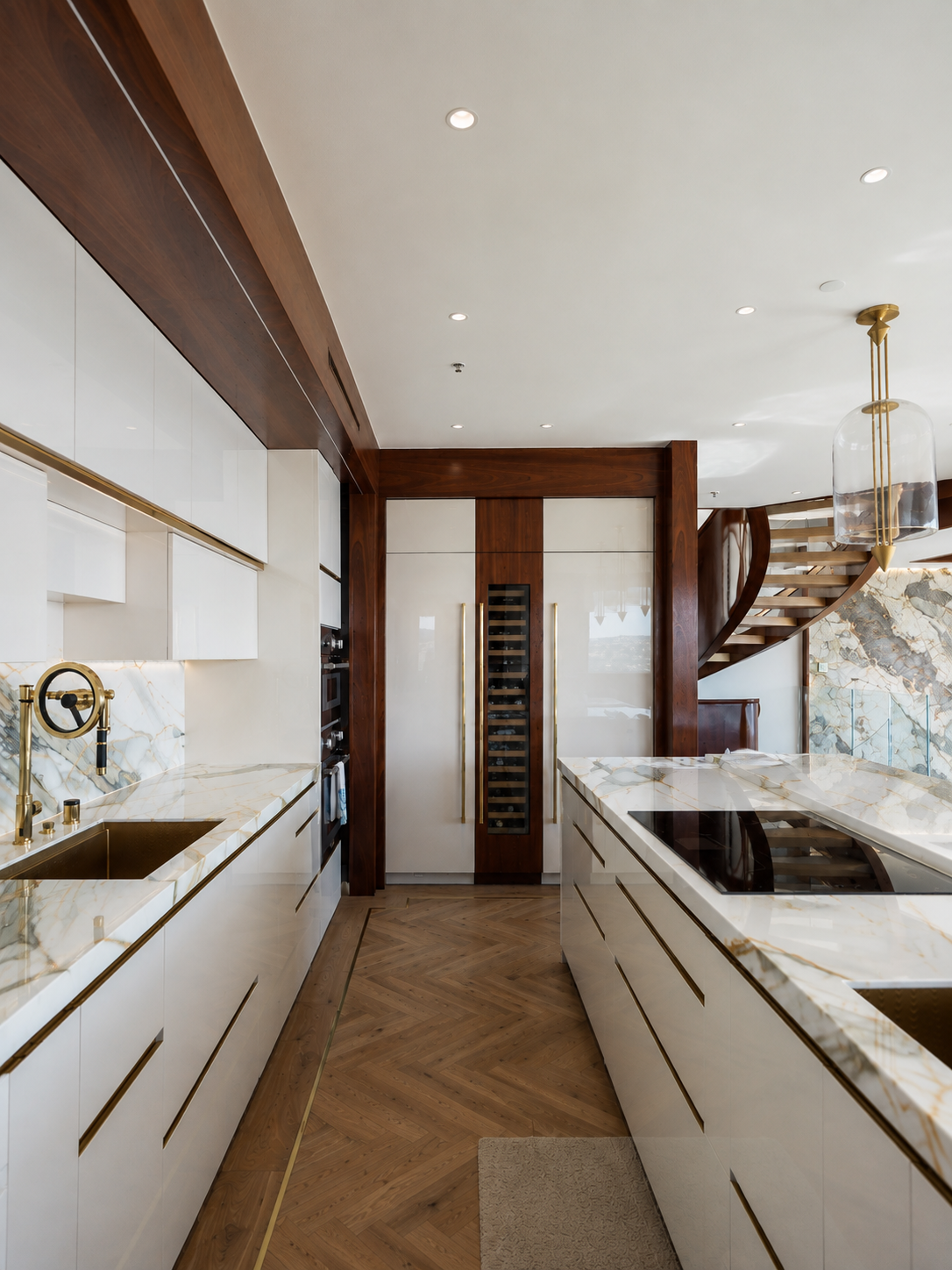 A luxury modern kitchen interior featuring white marble countertops, white cabinetry with gold handles, a dark wood herringbone floor, and a spiral staircase in the background.