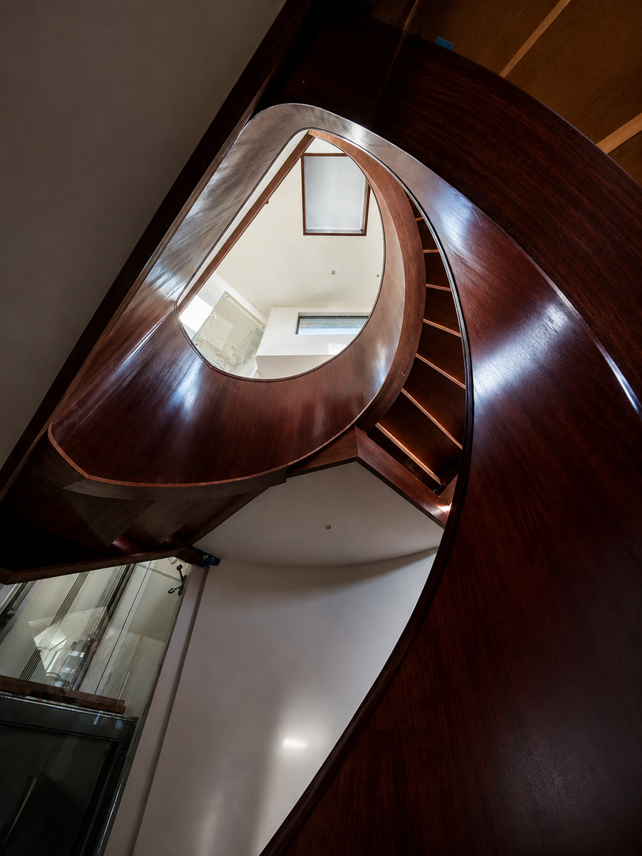 A dramatic low-angle view of a modern spiral staircase with a glossy dark brown mahogany wooden handrail and steps, overlooking a bright white interior atrium.